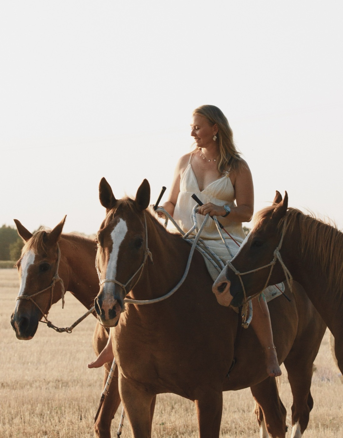 Woman in a white dress riding bareback among three chestnut horses